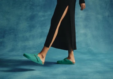 Model wearing green suede clogs and a black dress against a textured blue background for Birkenstock