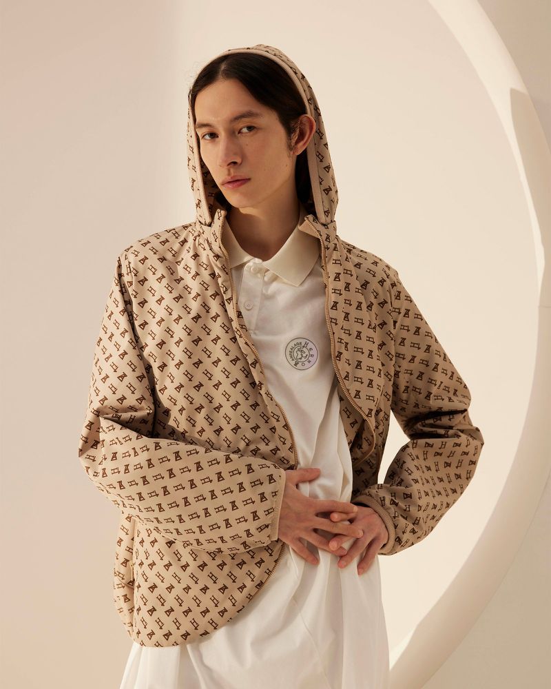 Young adult posing in a patterned beige hooded jacket and white polo shirt, standing indoors with soft, neutral lighting.