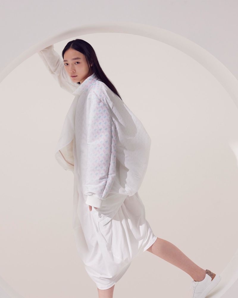 Fashion portrait of an Asian woman in white futuristic clothing, posing against a round white frame with white sneakers in a studio.