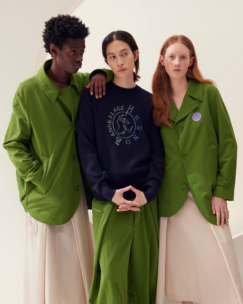 Three fashion models wearing jackets and long bottoms pose indoors against a light backdrop for a studio shot. The outfits are green and navy.