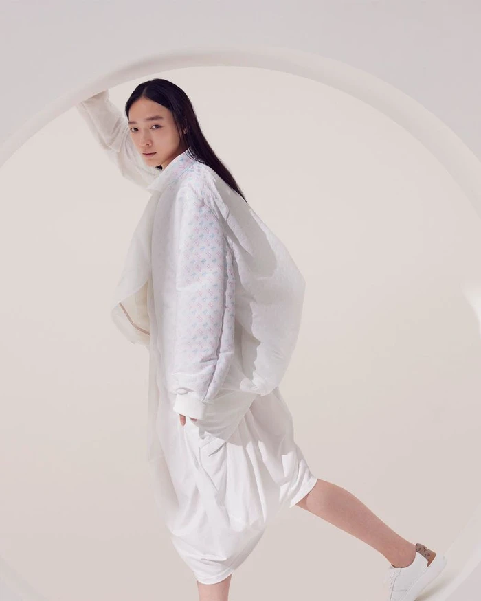 Model wearing white outerwear posing inside a curved architectural aperture against a seamless light background.