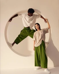 Two models posing inside a large circular wall cutout wearing Anrealage x Herno white shirts, green trousers, and white sneakers.