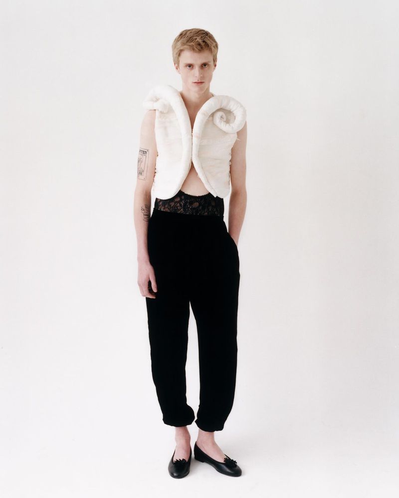 Young adult in a white sculptural top and black pants stands in a studio, showcasing a fashion-forward editorial look on a white backdrop.