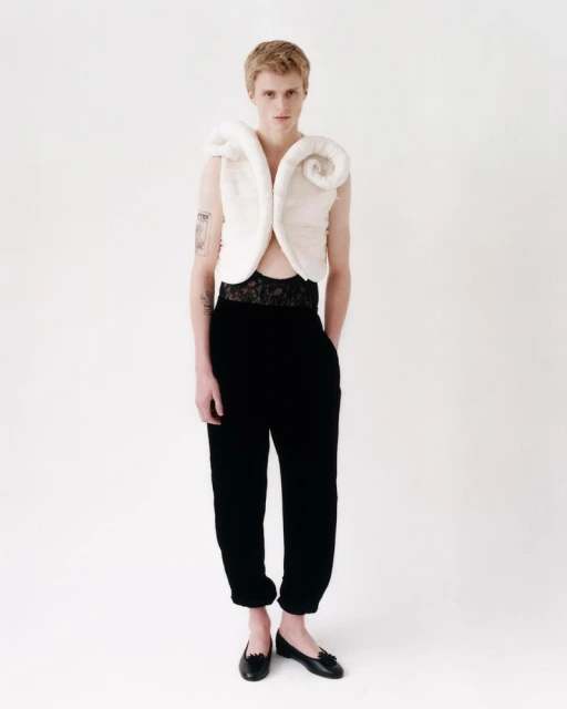 Young adult in a white sculptural top and black pants stands in a studio, showcasing a fashion-forward editorial look on a white backdrop.
