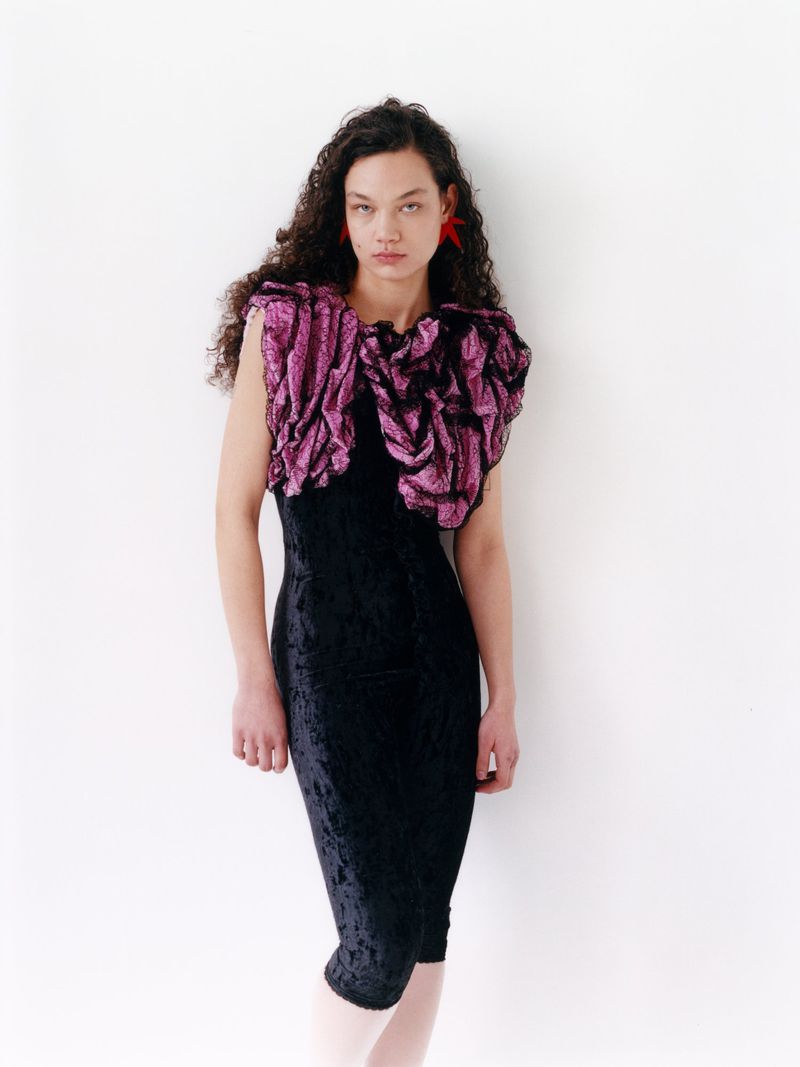 A woman stands in a photo studio in a black dress with a large pink ruffled top. She has curly brown hair and red star earrings.