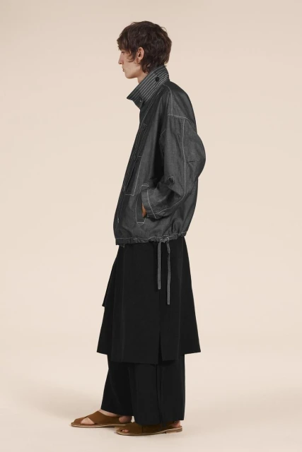 Profile shot of a non-binary fashion model in a dark grey jacket with contrast stitching and a long black skirt, against a peach backdrop.