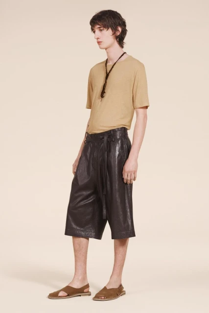 A light-skinned man models tan T-shirt, shiny dark brown culottes with tie, and sandals in a studio shot against a neutral backdrop.