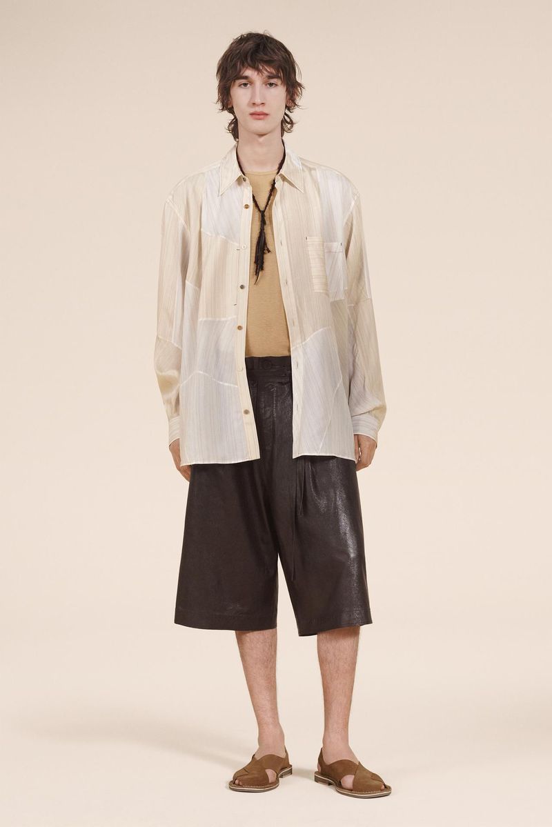 Full-shot of a male model wearing a tan short-sleeve shirt, a beige, patterned long-sleeve shirt, and brown sandals, posing in a studio.