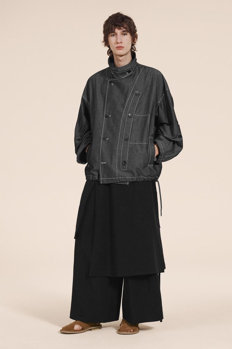 Male model in a dark denim jacket and black skirt poses in a full shot against a beige backdrop in a studio setting, fashion editorial.