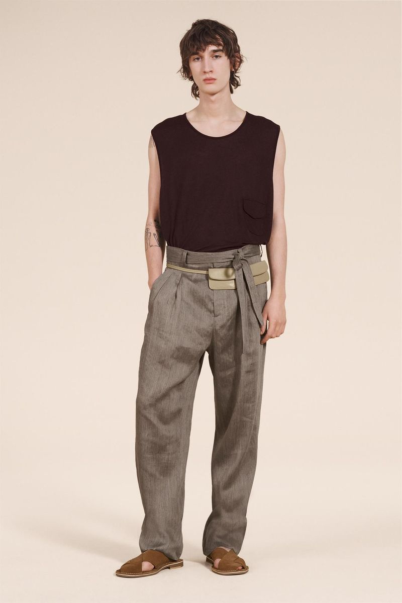 A slender man poses in a sleeveless dark top and pleated grey trousers with sandals, accessorized with a beige belt-bag, against a plain background.