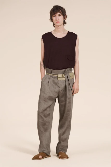 A slender man poses in a sleeveless dark top and pleated grey trousers with sandals, accessorized with a beige belt-bag, against a plain background.