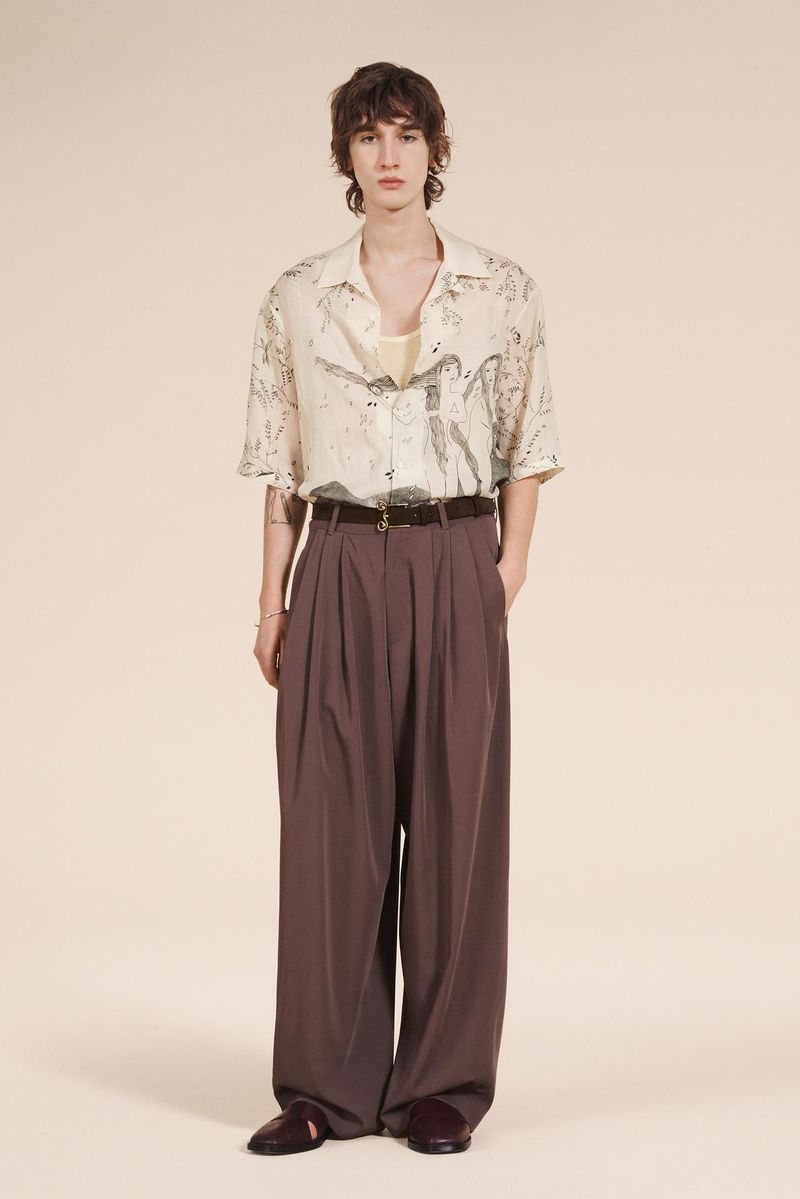 Standing male model in off-white patterned button-up shirt, wide leg brown pleated trousers, and burgundy shoes in studio.