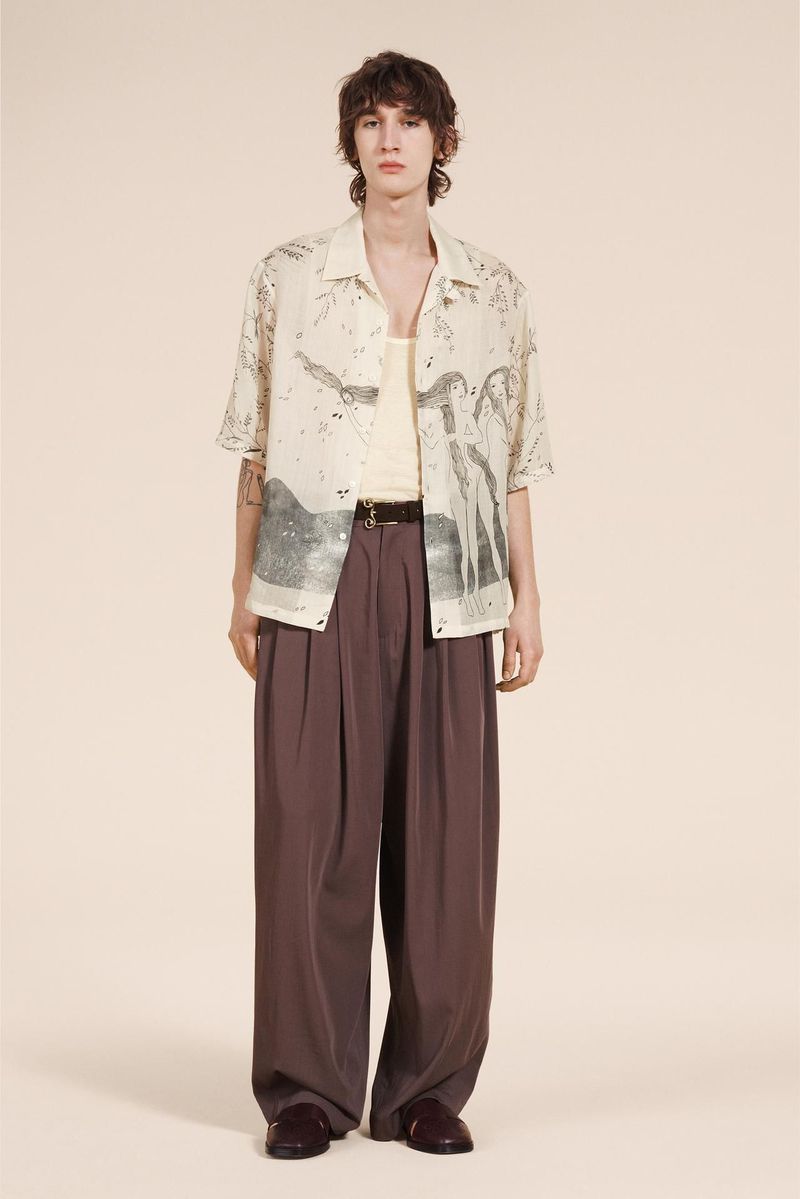 Full-length shot of a young adult male fashion model wearing a patterned short-sleeved shirt and baggy brown trousers in a studio setting.