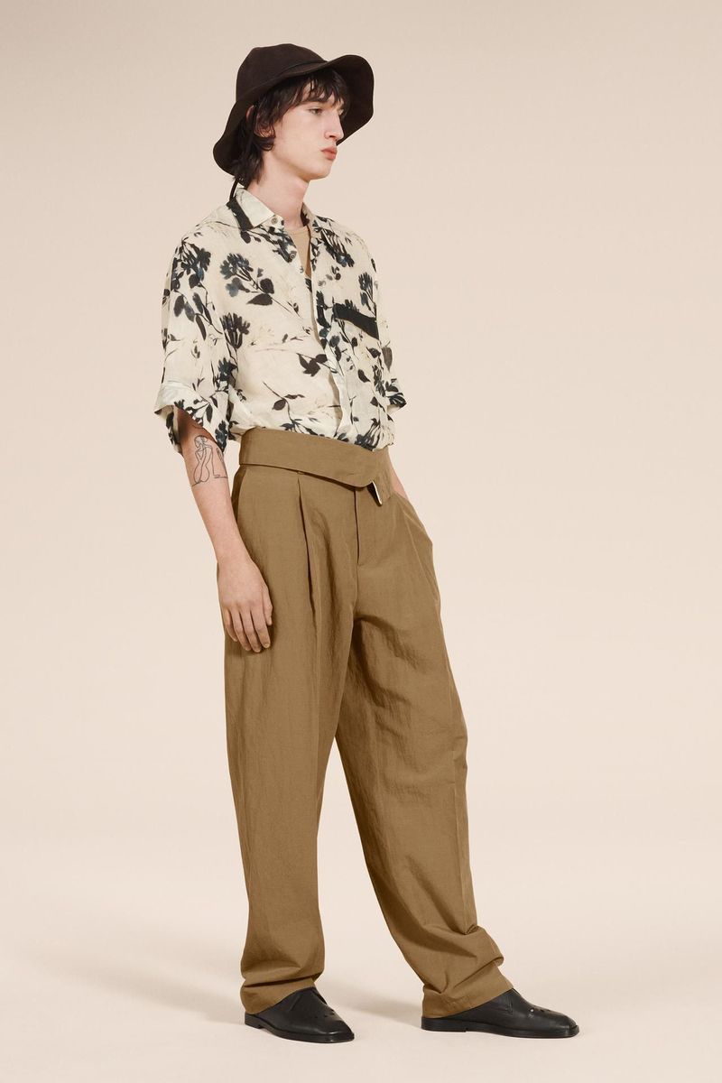A single adult male stands against a neutral background, modelling a short-sleeved floral shirt, wide-leg trousers, black shoes, and a bucket hat.