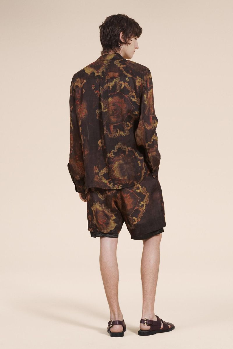 Man in dark, patterned shirt and shorts with brown sandals poses against a tan backdrop in a fashion or editorial style shot.