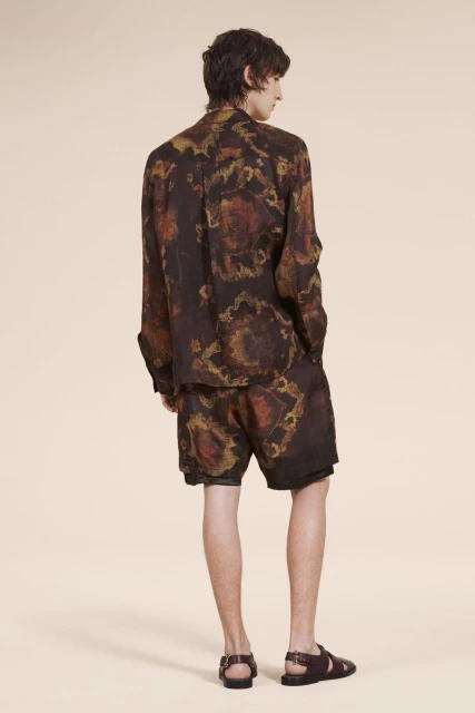 Man in dark, patterned shirt and shorts with brown sandals poses against a tan backdrop in a fashion or editorial style shot.