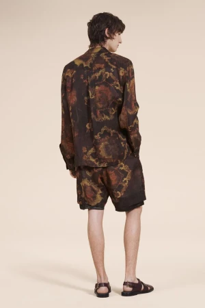 Man in dark, patterned shirt and shorts with brown sandals poses against a tan backdrop in a fashion or editorial style shot.