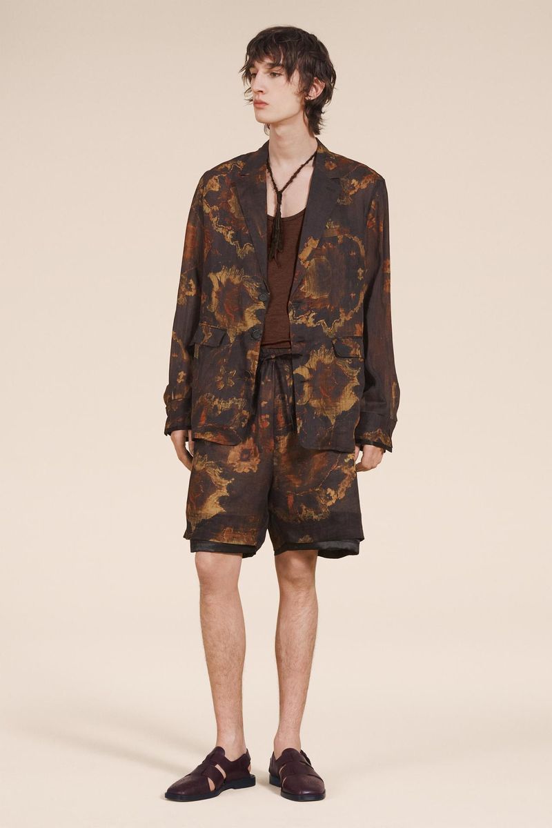 A young adult male fashion model poses in a brown and tan patterned jacket, shorts, and loafers against a neutral backdrop.