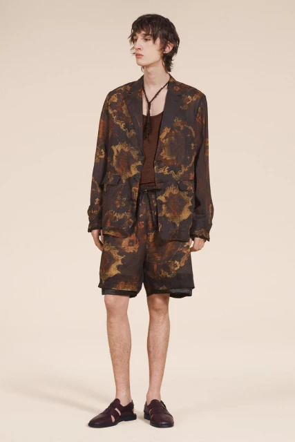 A young adult male fashion model poses in a brown and tan patterned jacket, shorts, and loafers against a neutral backdrop.