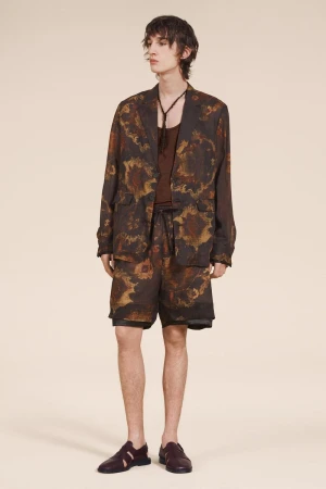 A young adult male fashion model poses in a brown and tan patterned jacket, shorts, and loafers against a neutral backdrop.