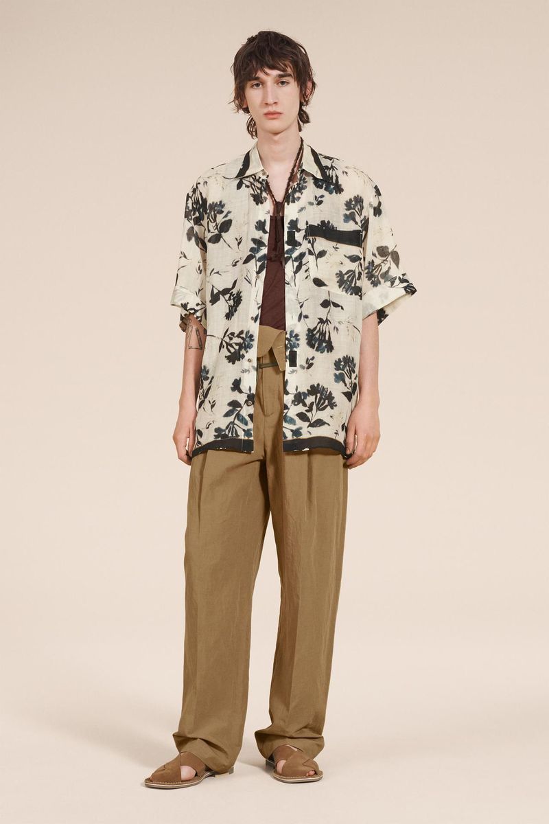 A stylish adult male in an open floral shirt and relaxed pants poses in a fashion editorial shot against a neutral backdrop.