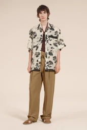 Model wearing a floral short-sleeve shirt, brown trousers, and sandals against a plain neutral studio background.