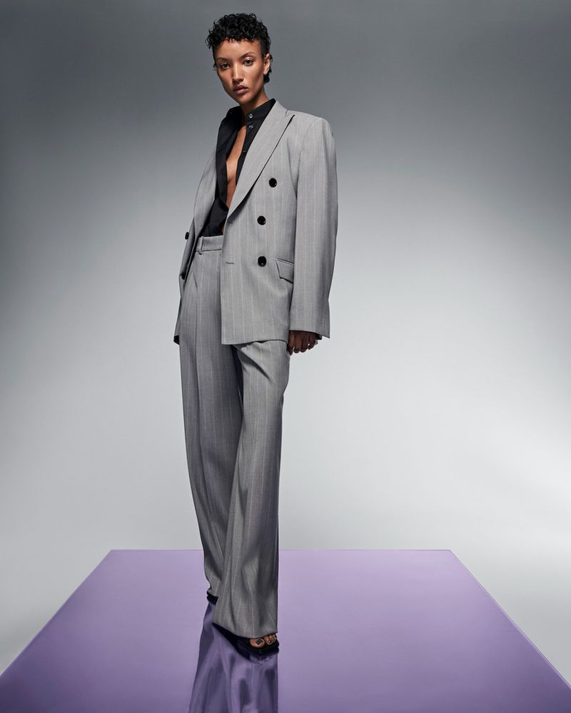 A model poses wearing a grey pinstripe suit with a black shirt on a reflective platform against a solid grey gradient background.