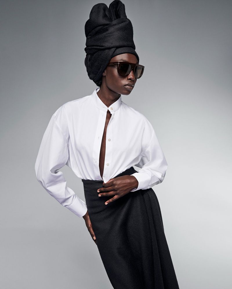 Dark-skinned woman in a white blouse, black skirt, and sunglasses stands against a neutral background, wearing a black head covering.