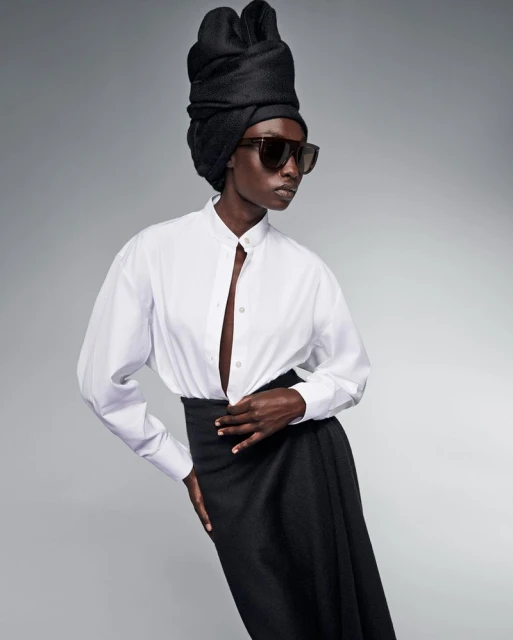 Dark-skinned woman in a white blouse, black skirt, and sunglasses stands against a neutral background, wearing a black head covering.