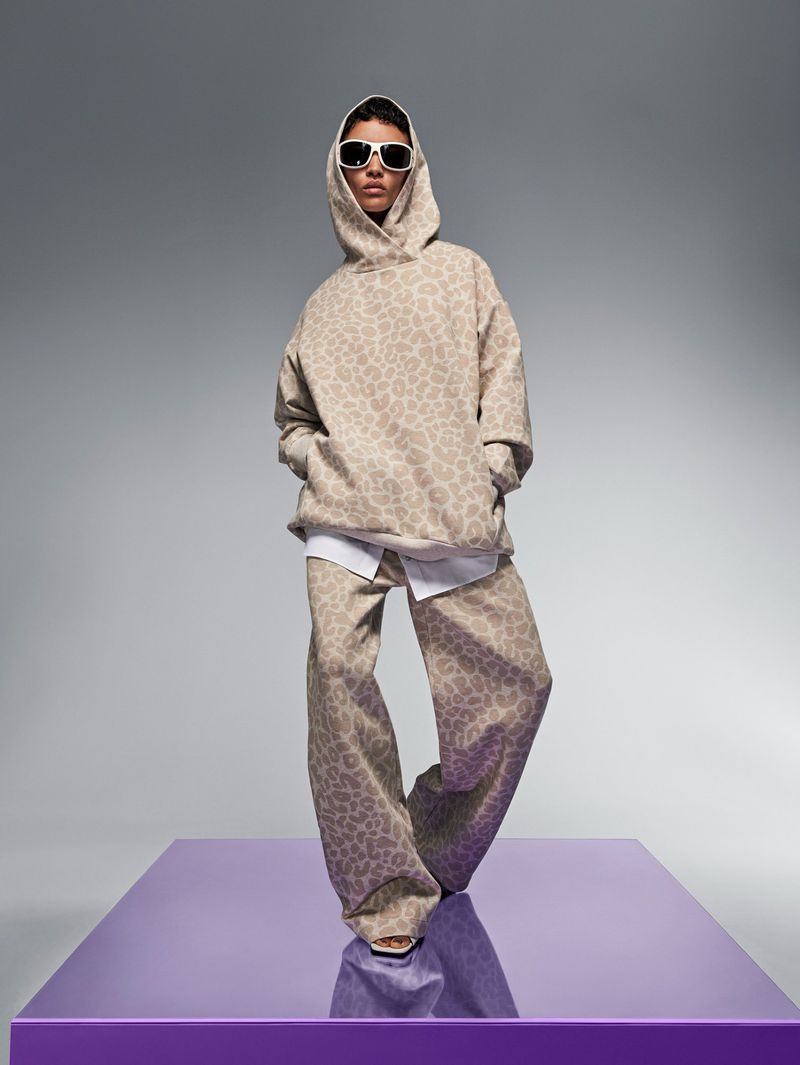 Fashion model poses wearing a beige patterned hoodie and pants, paired with white sunglasses, in a studio against a gray backdrop.