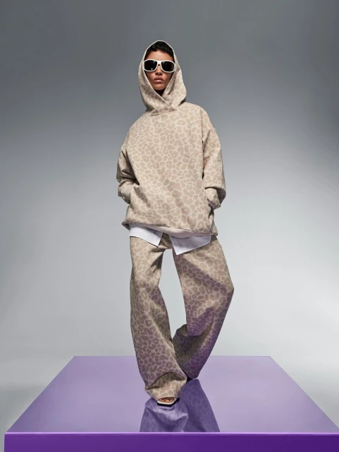 Fashion model poses wearing a beige patterned hoodie and pants, paired with white sunglasses, in a studio against a gray backdrop.