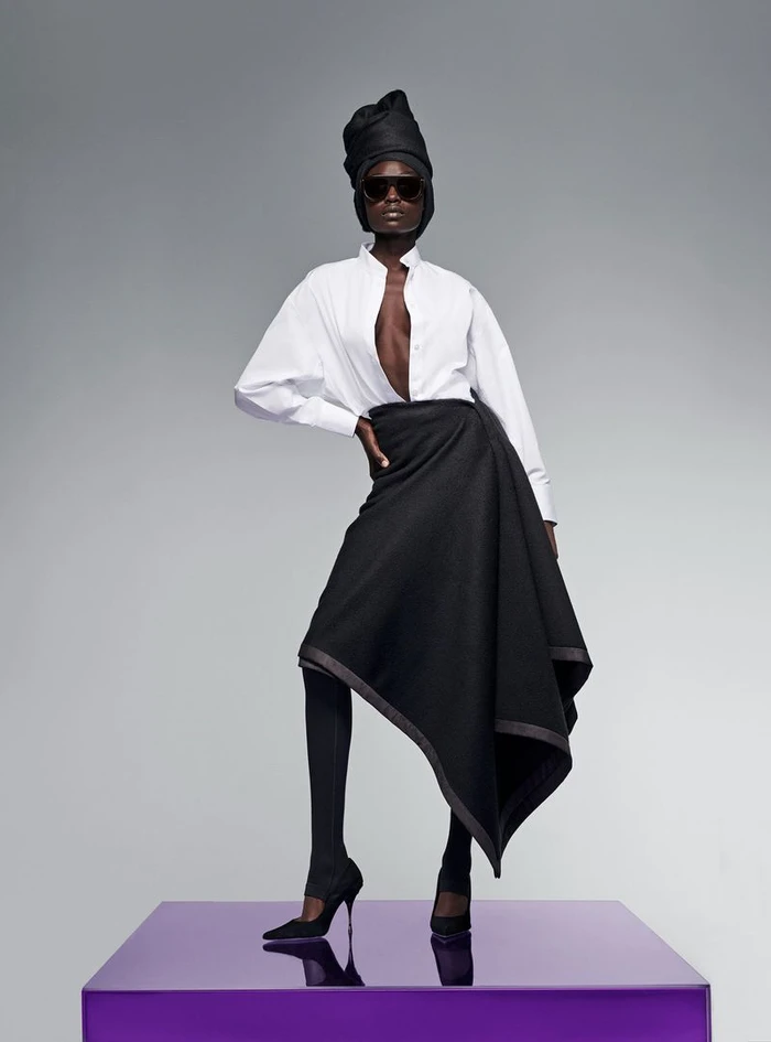 Model wearing a white shirt and black wrap skirt standing on a purple podium against a grey seamless background