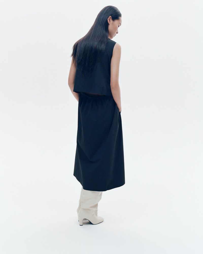 Back view of an adult woman in a black sleeveless top, black midi skirt, and light neutral ankle boots, posing against a white backdrop.