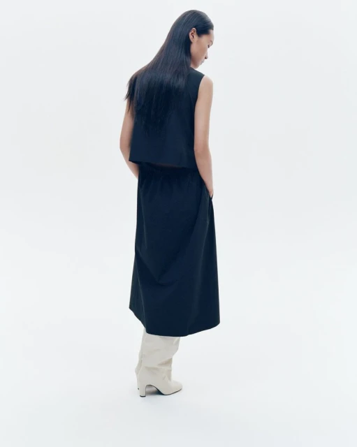 Back view of an adult woman in a black sleeveless top, black midi skirt, and light neutral ankle boots, posing against a white backdrop.