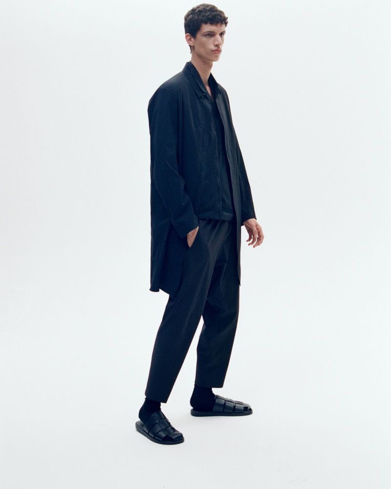 A young adult male poses in a black button-down shirt, black pants, black socks, and black sandals against a white backdrop.