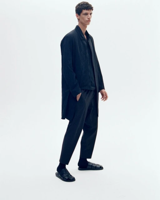 A young adult male poses in a black button-down shirt, black pants, black socks, and black sandals against a white backdrop.