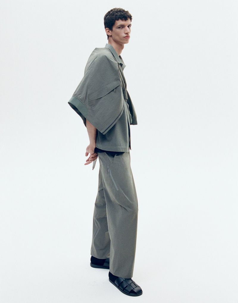 Full-length studio shot of a young adult man in an olive-green, oversized collared shirt and wide-leg pants, posing on a white backdrop.