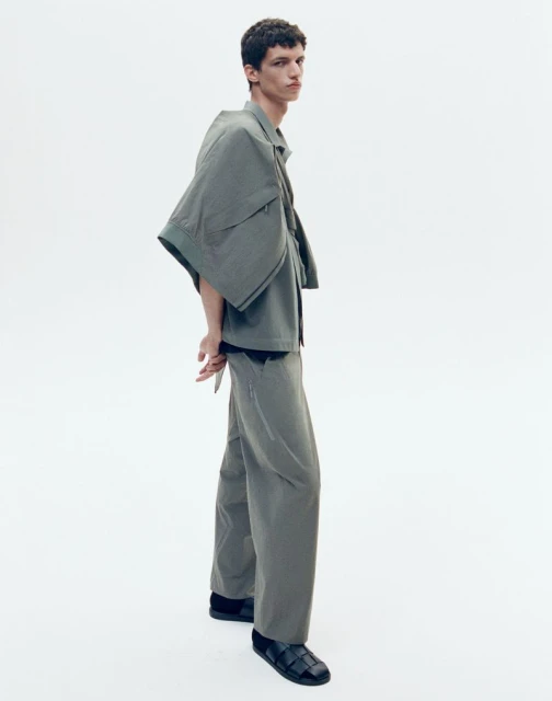 Full-length studio shot of a young adult man in an olive-green, oversized collared shirt and wide-leg pants, posing on a white backdrop.