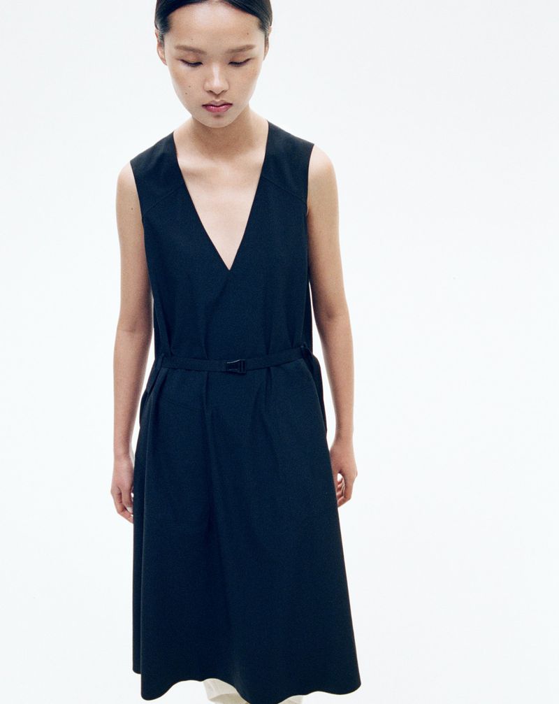 An Asian woman stands in a photo studio, wearing a sleeveless black dress with a V-neck and a belt, against a plain white backdrop.