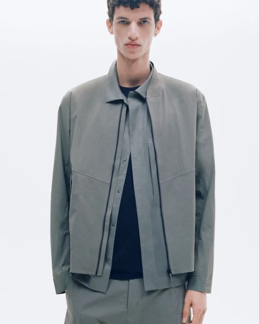 Studio shot of a young adult male model wearing a grey collared jacket with dark shirt and matching pants against a white background.