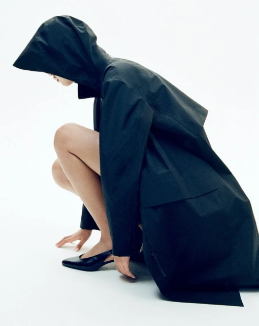 A woman in a hooded black coat squats against a white background, looking down, with a single black heel visible.