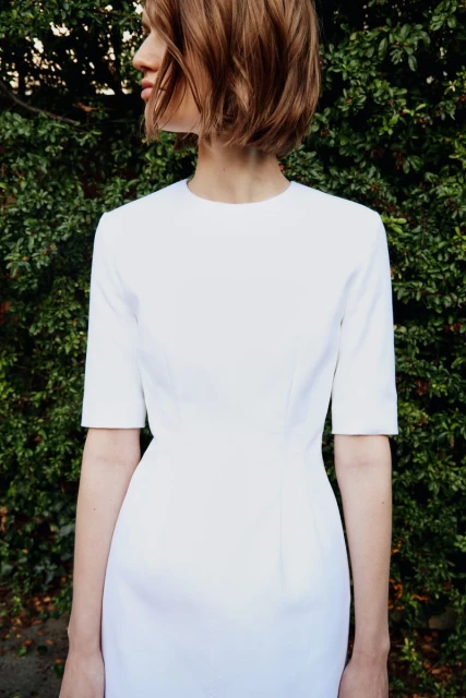 A woman with short brown hair models a white dress outdoors with a green foliage background. The dress is fitted and simple.
