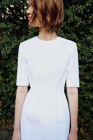 A woman with short brown hair models a white dress outdoors with a green foliage background. The dress is fitted and simple.
