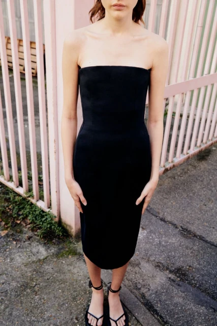 A woman stands outside in a black strapless dress and strappy sandals in front of a pale pink fence on a concrete surface.