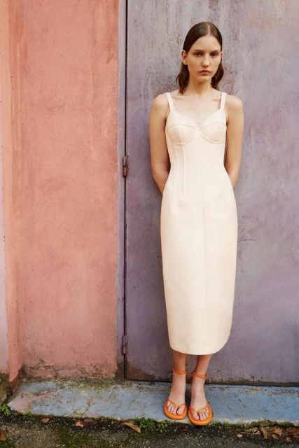 Woman in a cream-colored dress with spaghetti straps stands against a weathered door, complemented by her orange sandals and neutral makeup.