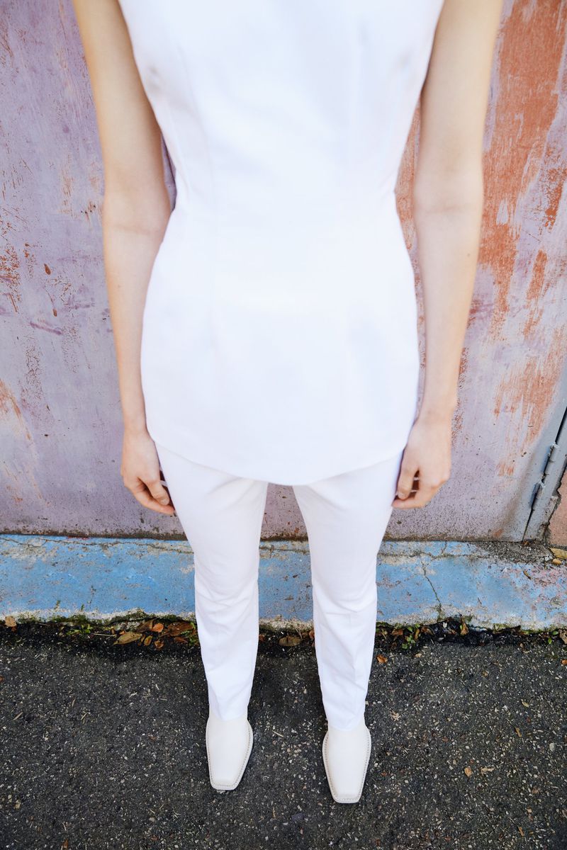 A woman in a white outfit stands against a textured wall. The photo emphasizes the outfit and the contrast between the garment and wall.