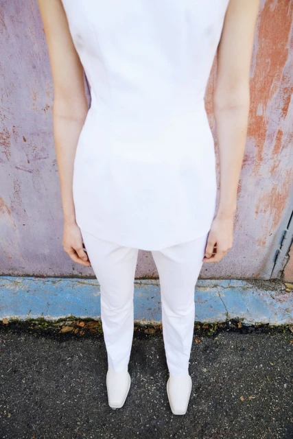 A woman in a white outfit stands against a textured wall. The photo emphasizes the outfit and the contrast between the garment and wall.
