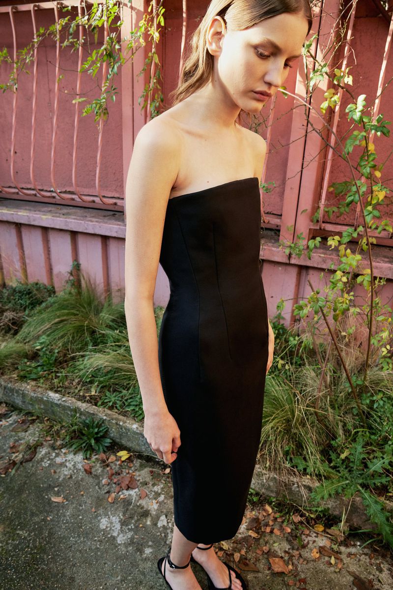 A woman in a black strapless dress stands outdoors with greenery and a pink wall visible in the background, captured in natural light.