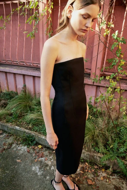 A woman in a black strapless dress stands outdoors with greenery and a pink wall visible in the background, captured in natural light.