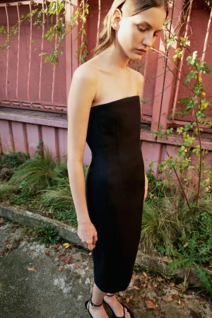 A woman in a black strapless dress stands outdoors with greenery and a pink wall visible in the background, captured in natural light.